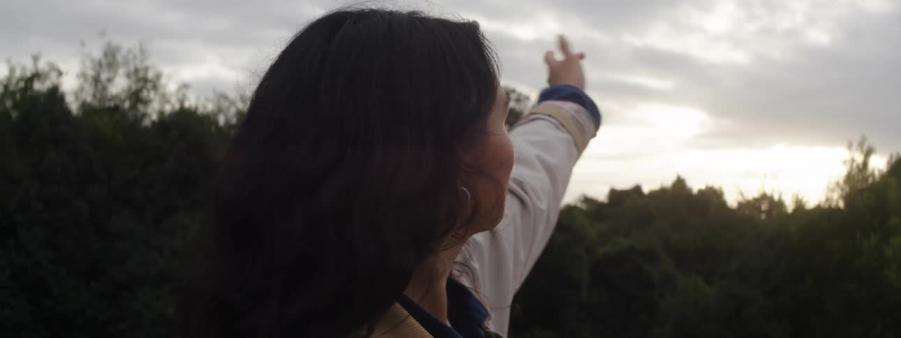 Woman with black hair hoop earrings and white jacket lifts her hand up against the sunset in the forested area