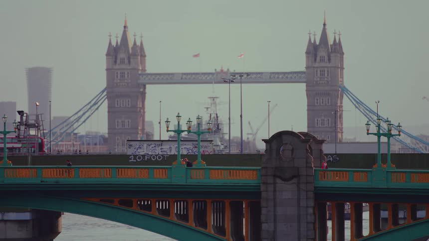People walk across a bridge in front of a double decker bus driving pas tall buildings on the other side of the bridge