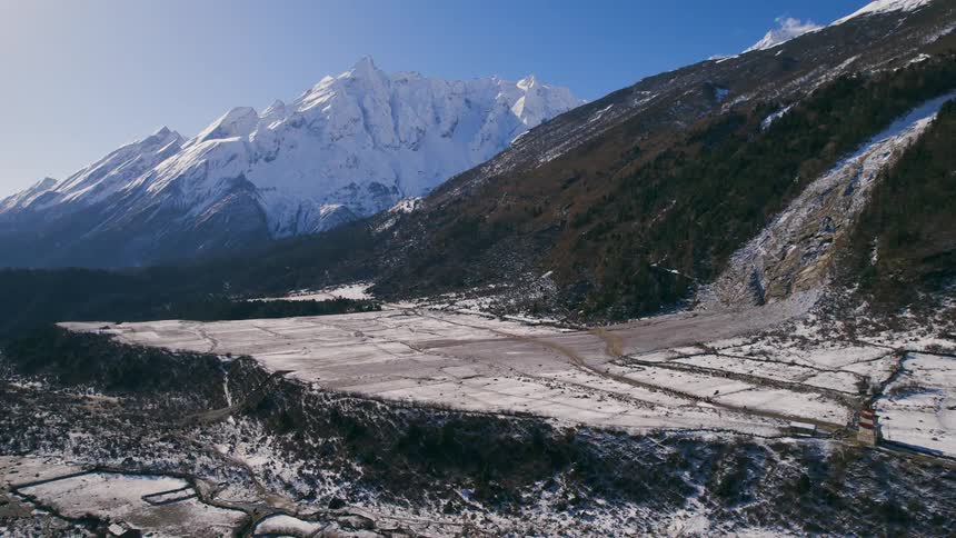 An aerial of flatlands at the bottom of a mountain with snow covered mountain peaks in the distance
