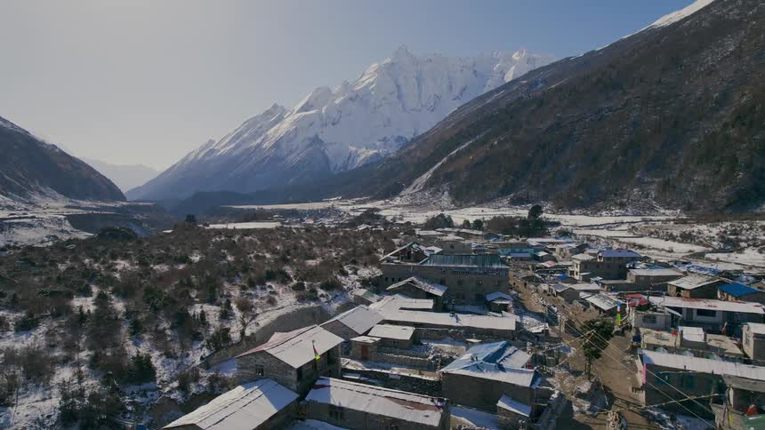 An aerial of a rural village in a valley between mountains with snow covered peaks in the middle of the day
