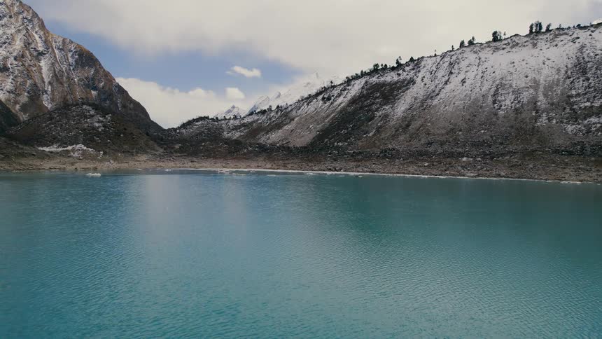 An aerial of a turquoise lake surrounded by snow covered hillsides on a bright but cloudy day