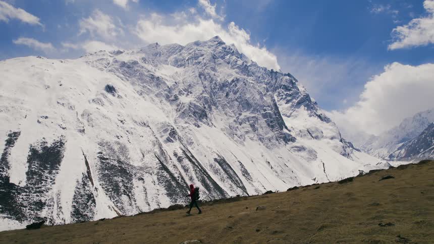 Hiker with red coat backpack and poles walks downhill with snowy high mountain ridge in the background