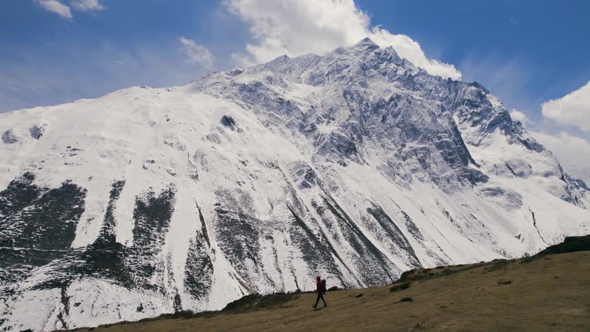 A man wearing a red hoodie and a backpack is walking across a mountain top holding trekking poles overlooking a snow covered mountain