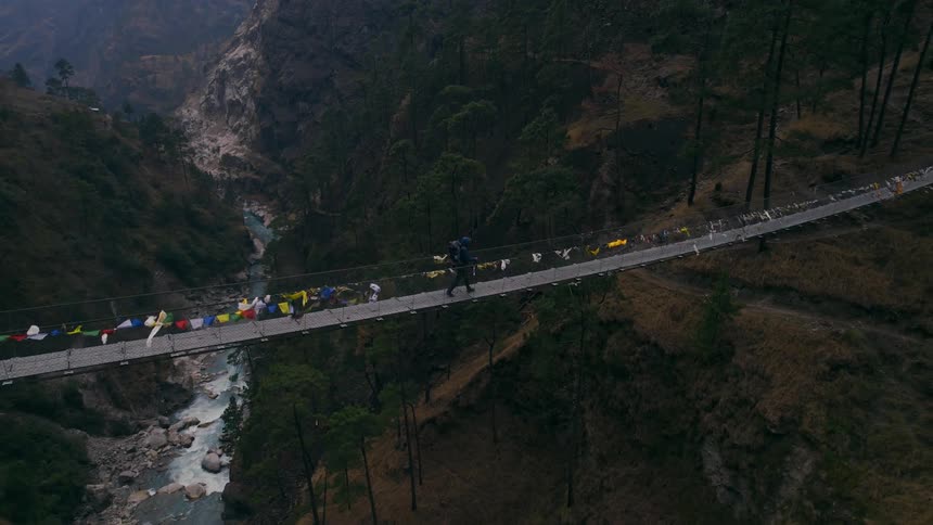 An aerial of a person walking over a suspension bridge going over a ravine between two mountains