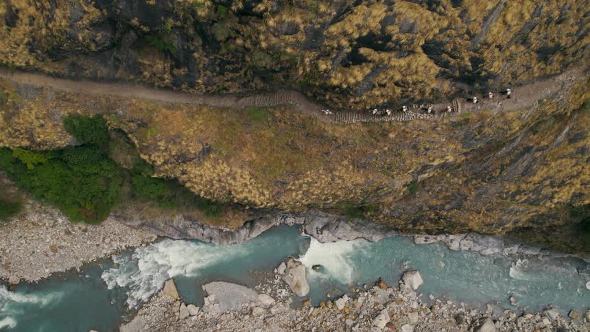 The aerial view captures a group of people walking along a dirt track on a mountain ridge with a river flowing gracefully beside them