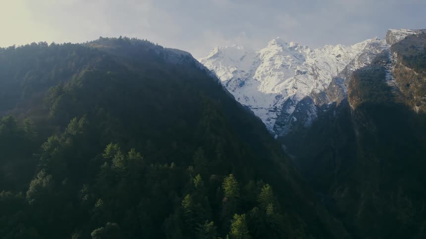A dramatic zoom of snow covered mountain peaks and green forests on the hillside