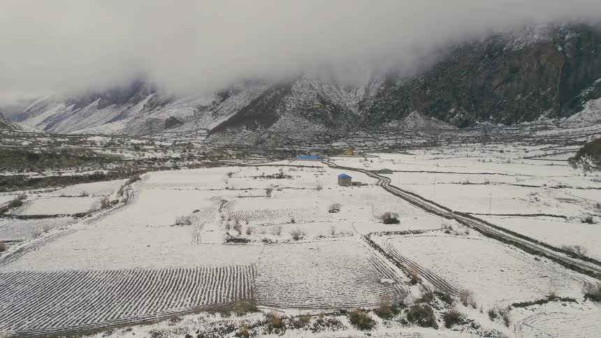 An aerial of snow covered lands with a dirt road across the landscape connecting houses with cloud covered mountains in the distance