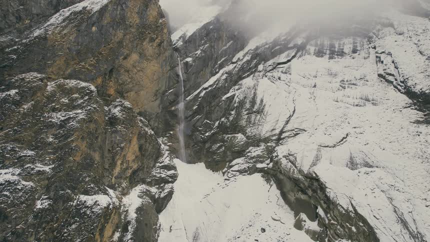 A narrow waterfall is nestled within a rocky mountain range covered in snow
