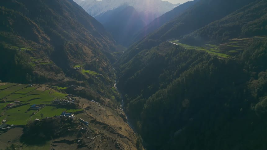 A mountain range with trees a river a few houses and fields used for agriculture