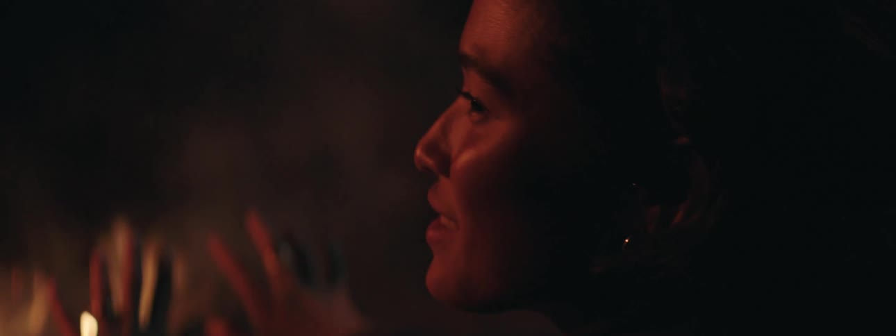 A woman at a campfire at night engaged in conversation while spreading her fingers out to get warm
