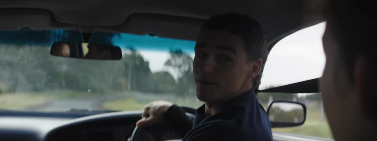 A man driving a car down a rural road turning around looking at a person in the seat behind him while they talk