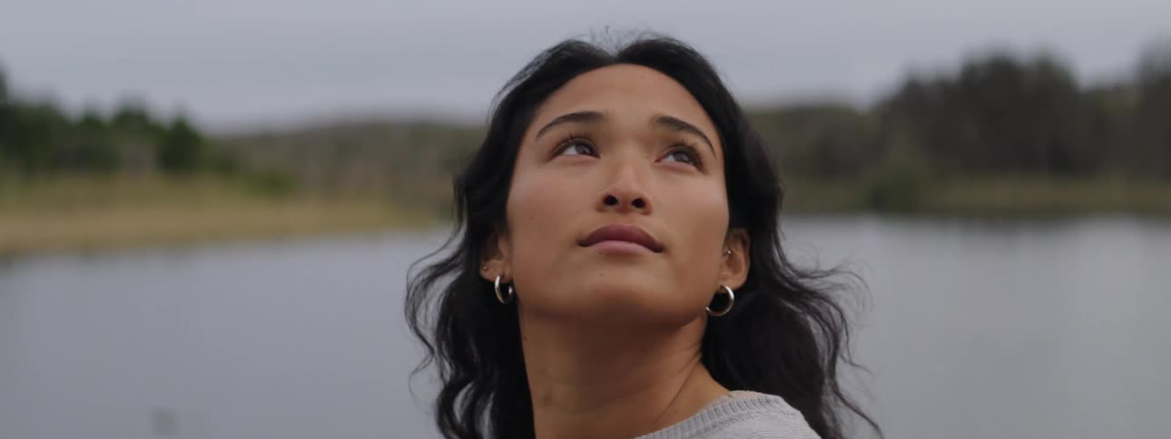 Woman with dark wavy hair and earrings looks over her shoulder by lake