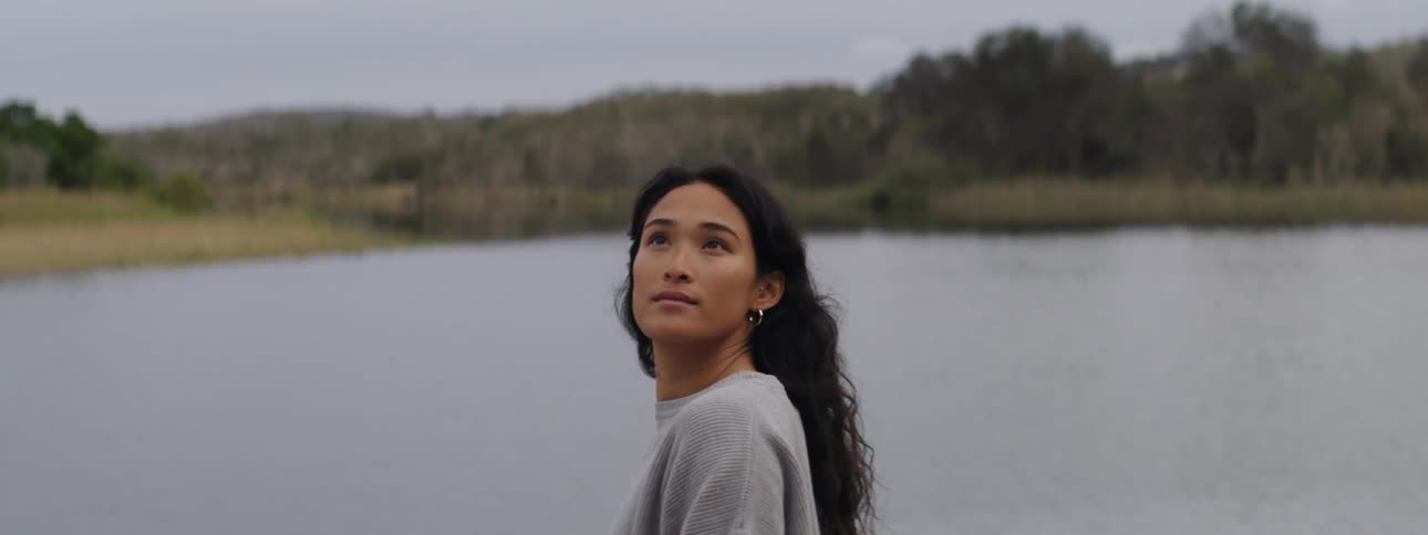 Woman standing on lake shore across from hills and woods looks around with a slight smile