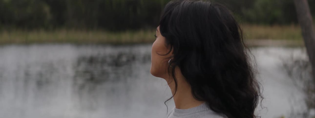 A young woman stands near a river as the wind blows her hair