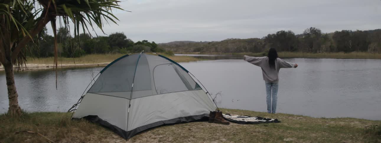 A woman getting out of her tent walking over to a waters edge while stretching and looking around
