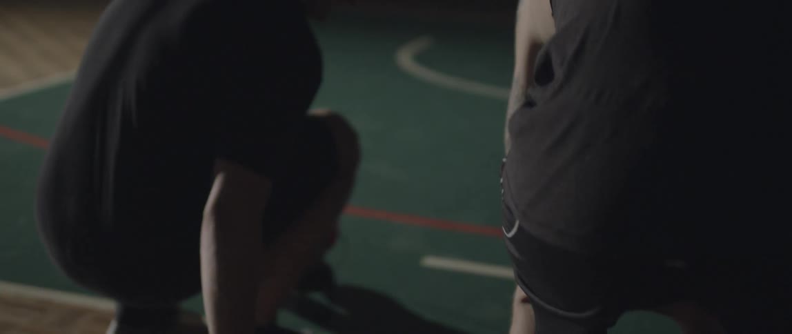 Two men carrying athletic shoes sit down on a court and put their shoes on