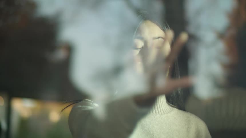 A young woman with her eyes closed holds her hand towards a window and then brings it up to her face