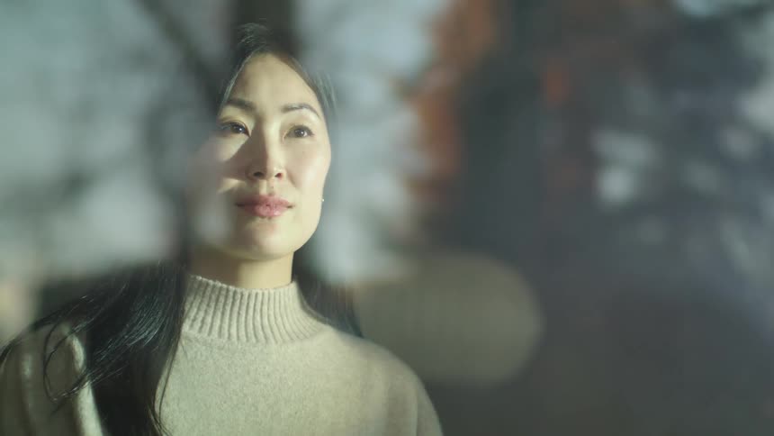 A woman stands before a window with her eyes closed for a moment before opening them and gazing outside