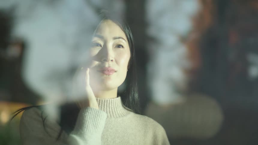 A woman standing next to a window with the reflection of trees the sky and clouds
