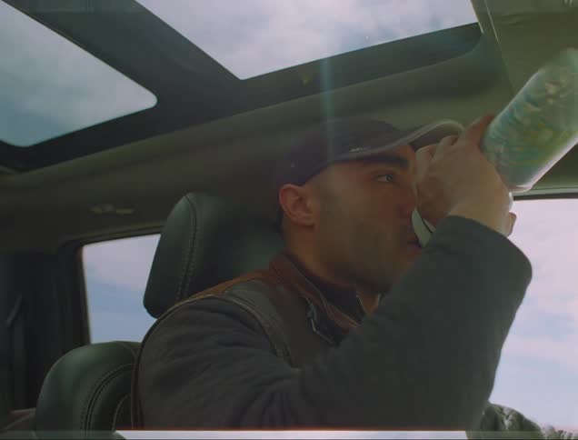 The sunroof over his head shows a cloudy sky as driver takes a drink from water bottle