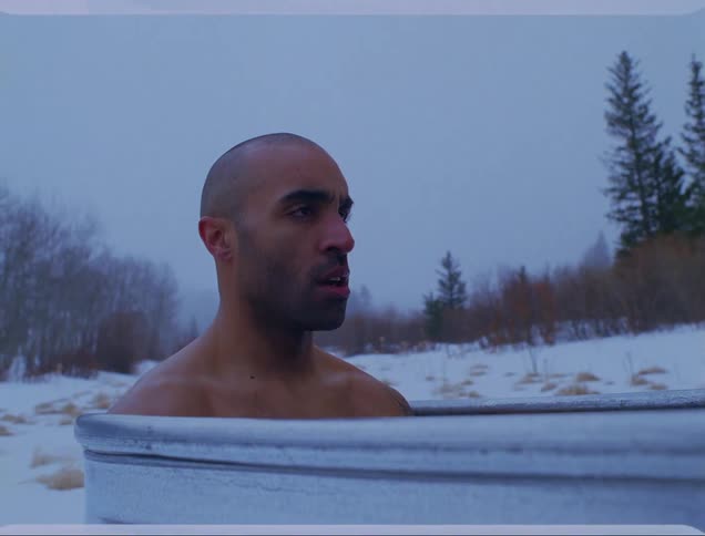 A man soaks in a metal tub outdoors in winter near a forest