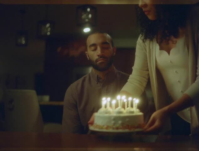 Man sits at table looking sad while woman carries in birthday cake and sets in down in front of him and smiles for a second when she touches his arm then goes back to frowning