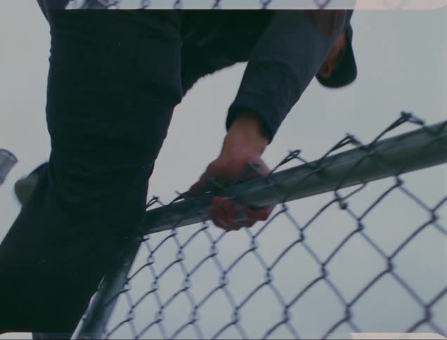 A man wearing a baseball cap leaps over a chain linked fenced outside on an overcast day
