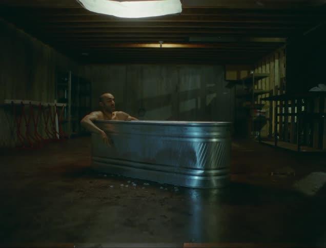 A man shivers as he soaks in a metal bathtub in an empty barn