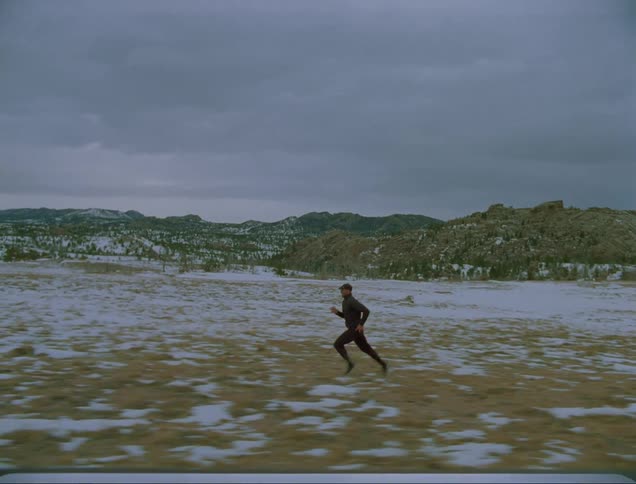 Man with a cap a black coat and jogging pants runs on snow covered plains on a cloudy day