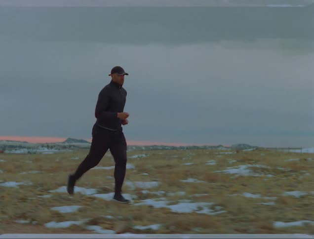 Man with a cap a black coat and jogging pants runs on snow covered plains on a cloudy day