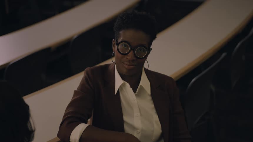 Black person wearing a red suit sitting down indoors turning their faces and staring at the shot