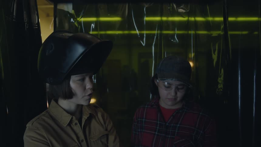 Two women standing in a plastic covered room one wearing a helmet the other safety glasses