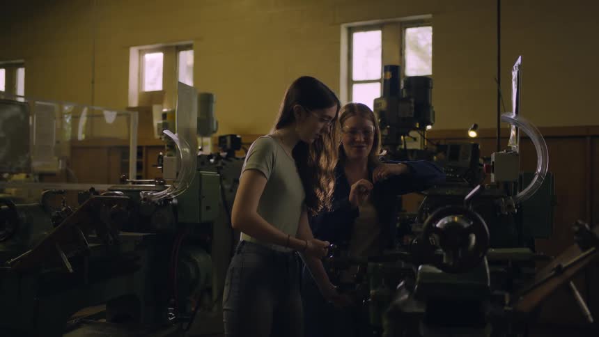 Young girls standing together near machinery as one turns a knob on the machine