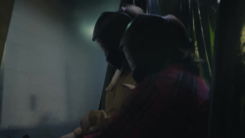 Two women put on protective welding gear while standing in dimly lit workshop