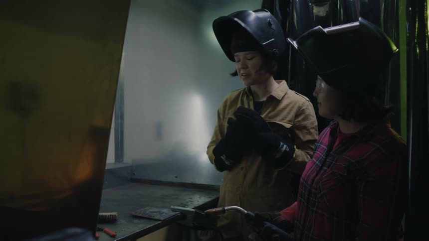 Two women in a room wearing gloves and headgear while welding