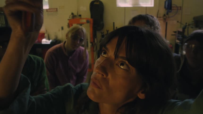 People watching a female mechanic work on a car with a screwdriver