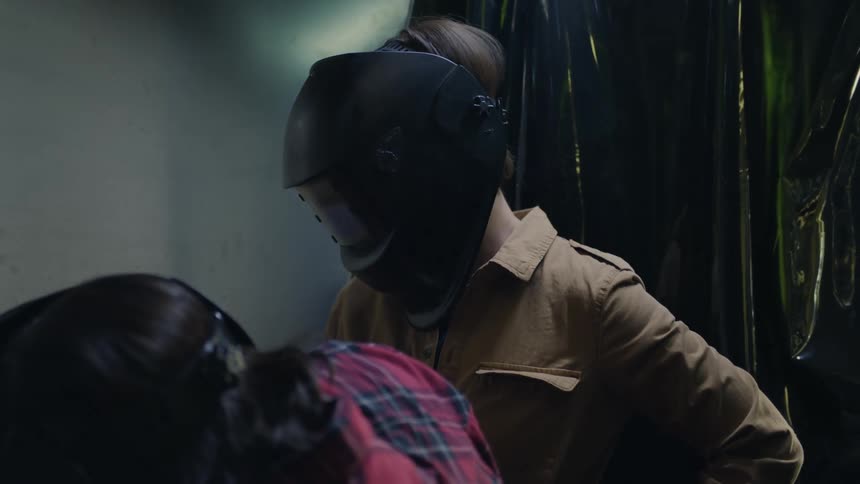 Two women wear welding masks in workshop as one of them welds