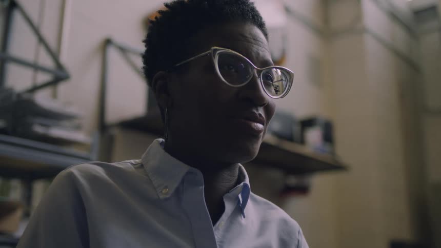 Short haired woman wearing glasses talks while sitting on a workshop