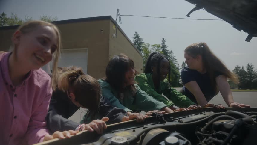 Five women pushing a vehicle with its hood open coming to a stop and celebrating by highfiving each other in jubilation