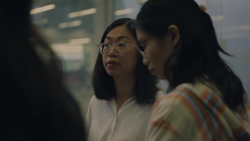 Two asian female standing in front of a large glass interior window with a lot of office lights reflecting from it are standing in a group discussing a project
