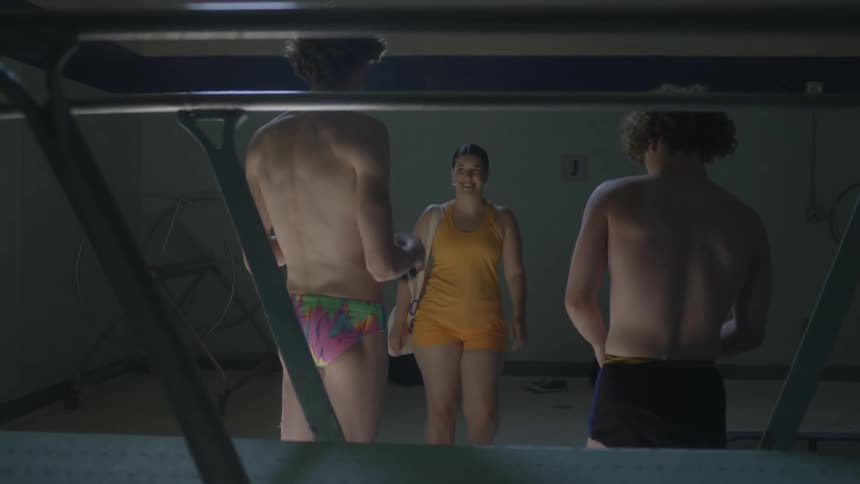 Woman in yellow bathing suit walking and then talking to two men who are preparing to go swimming