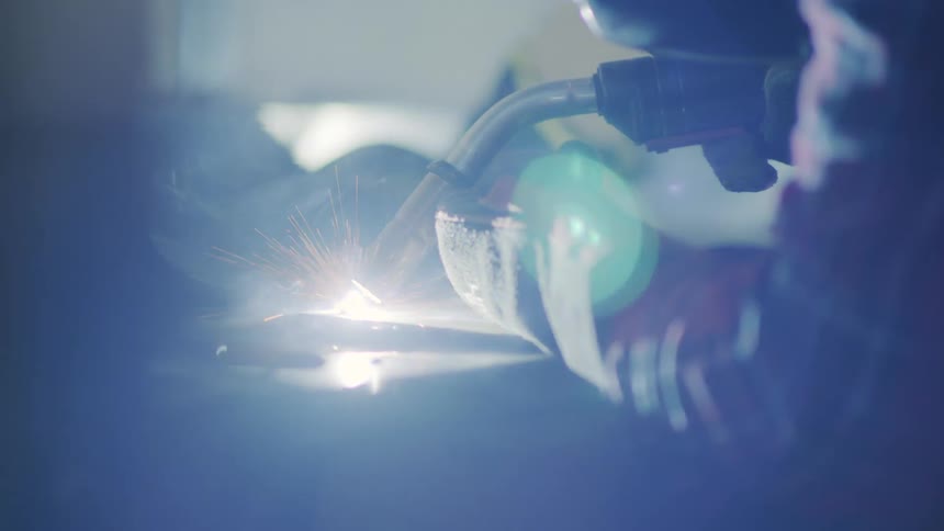 A welding torch sparking as it lights up and a person wearing protective gloves and face shield holds the torch over a piece of metal welding it over a table