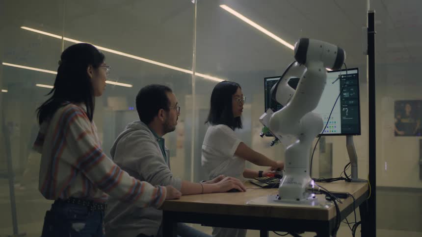 A trio of people discuss the usage of a computer controlled robotic arm