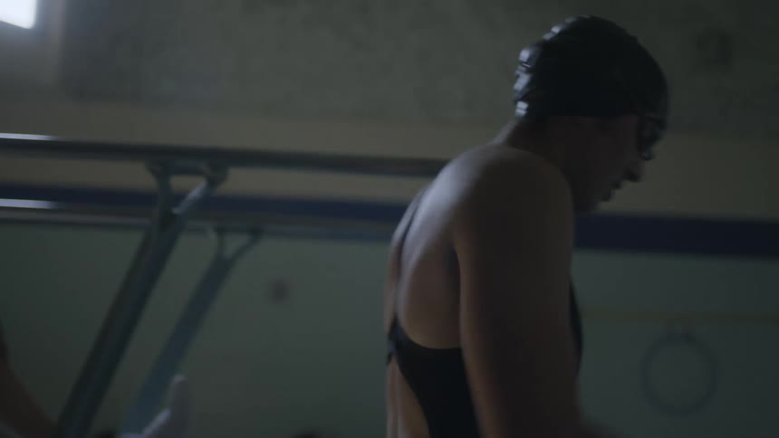 A woman in a swimsuit crouches under a diving board and dives into an indoor pool