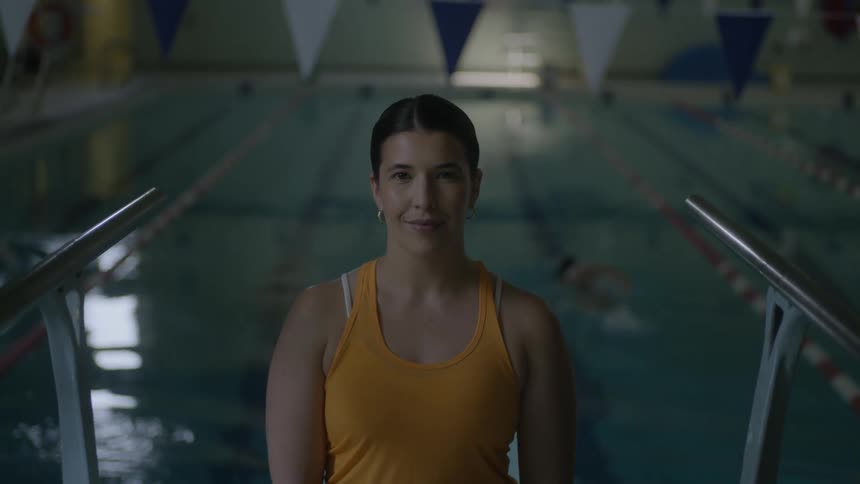 Swimmers in an indoor pool swim behind a woman in a tanktop