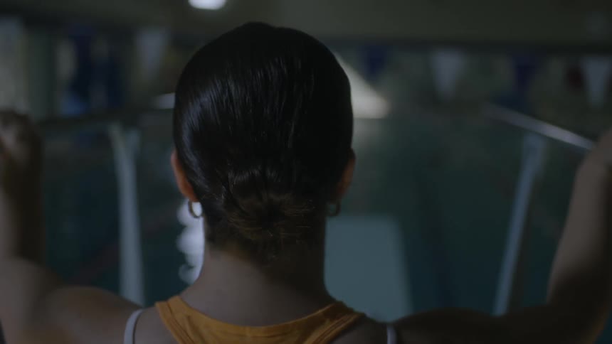 A woman grasping the handrail of a springboard leading to a swimming pool with the room enveloped in dim lighting