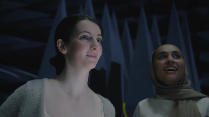 One woman consults computer screen as she and another woman look happily around industrial lab