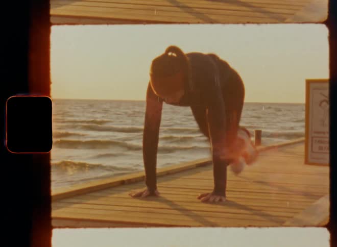 Attractive female athlete working out and doing mountain climbers on pier over the sea as the morning sun rises and the waves wash onto the beach during sunrise on vintage Super 8mm film