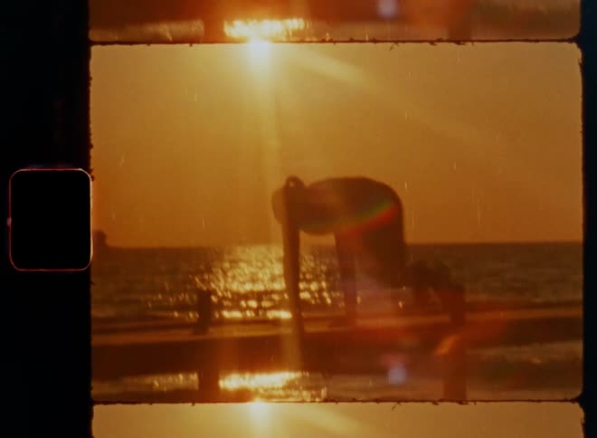 Silhouette of unrecognisable sporty woman doing mountain climbers for exhausting Pilates workout with fast heartbeat on wooden deck at the coast in fresh breeze surrounded by waves and golden sunlight with dynamic shaky Super 8mm camera movement