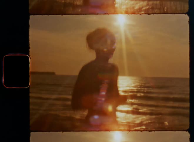 Dynamic handheld 8mm film of a young sporty woman running near the ocean as the sun is setting on the horizon over the sea with a red sky and lens flare